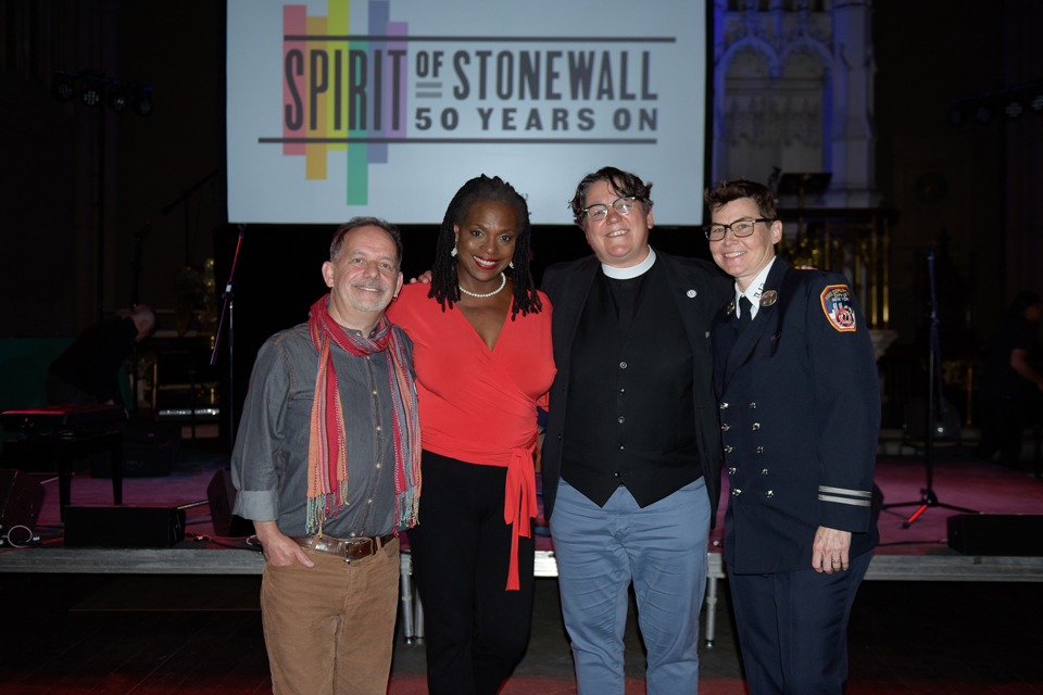 Four people standing in front of the "Spirit of Stonewall - 50 Years On" Banner at St. Ann and the Holy Trinity Pro Cathedral